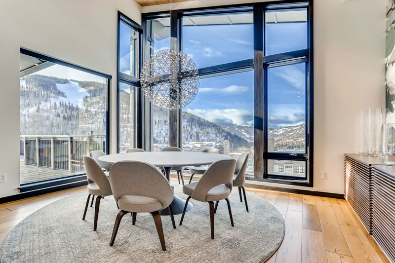A dining area with a round table, chandelier, and large windows showing a mountain view.